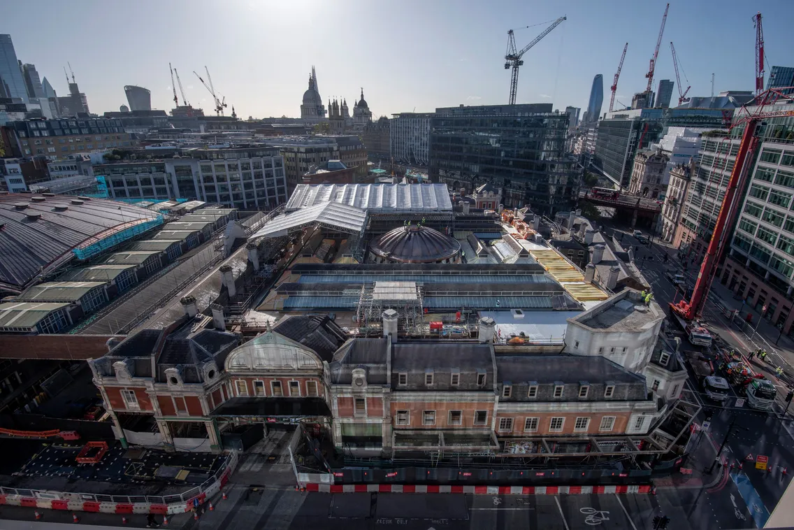 Aerial view of a city construction site surrounded by cranes and modern buildings, with a mix of older architecture in the foreground and a clear blue sky overhead.