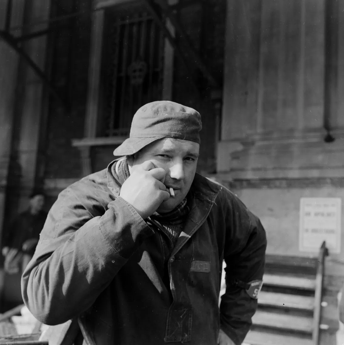 A black-and-white image of a man wearing a cap and work clothes, smoking a cigarette, standing in front of a building with architectural details.