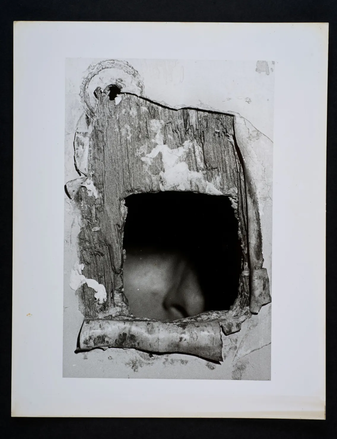 A black and white photograph shows a person's face partially visible through a small, rough hole in a deteriorating wall.