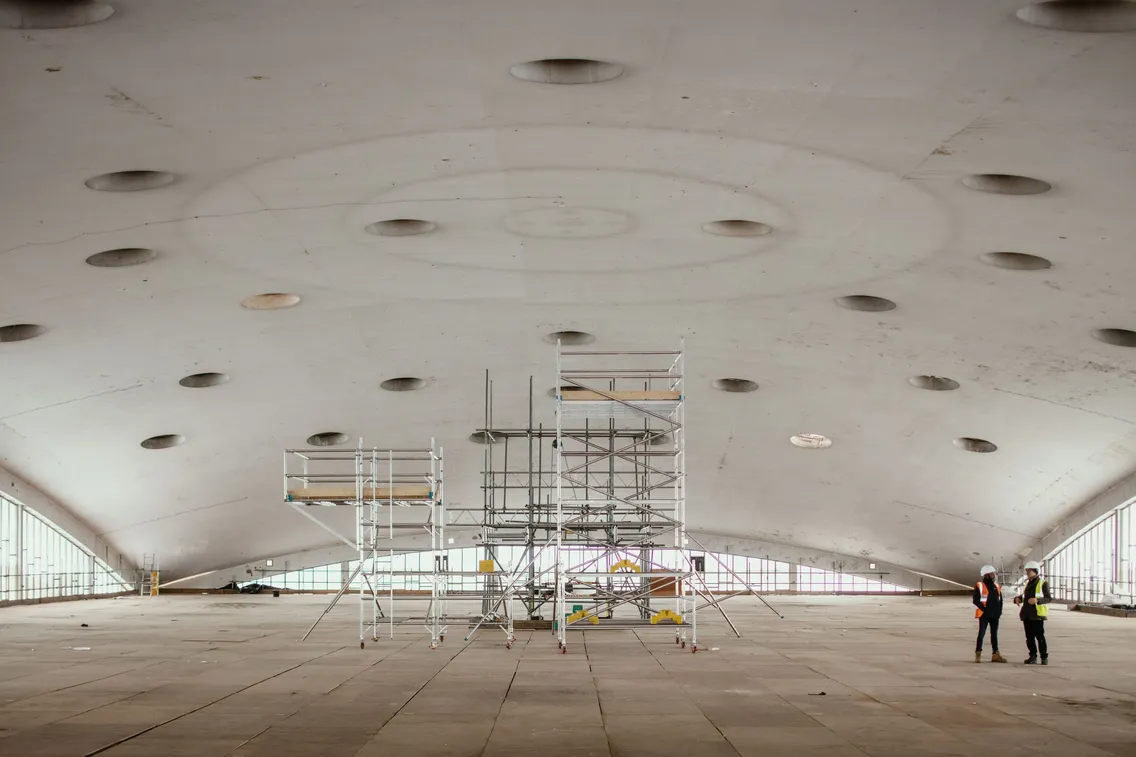 Large, unfinished building interior with a curved ceiling and numerous circular openings. Scaffolding is positioned in the center, and two workers in safety gear are standing to the right.