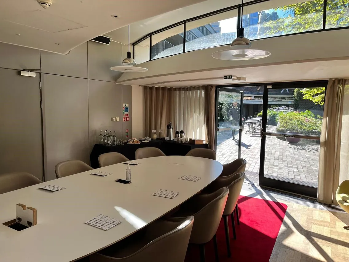 A modern conference room with a large oval table, chairs, and a red carpet leading to a glass door. Natural light streams in through the windows. A beverage station is set up in the corner.