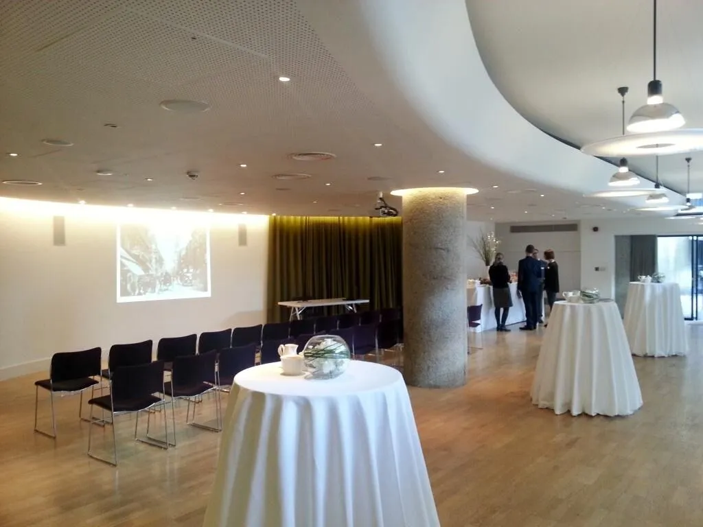 A modern conference room with round tables covered in white tablecloths, chairs arranged in rows, and people conversing near a green curtain. A projection screen is visible in the background.
