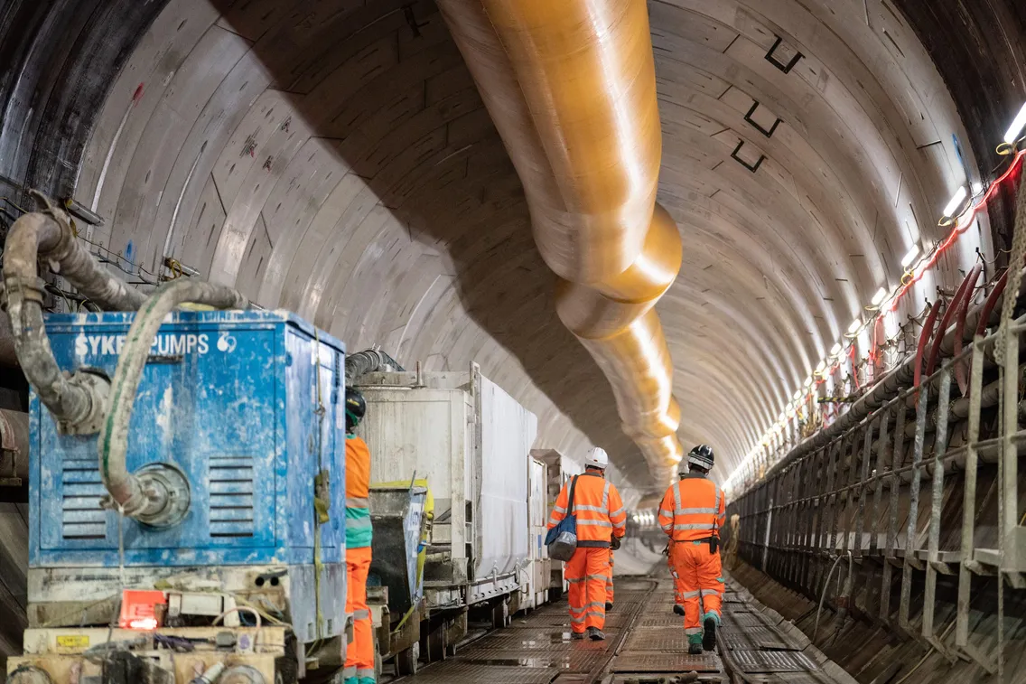Two construction workers in orange safety gear walk through an underground tunnel under construction, with large machinery and equipment visible on both sides.