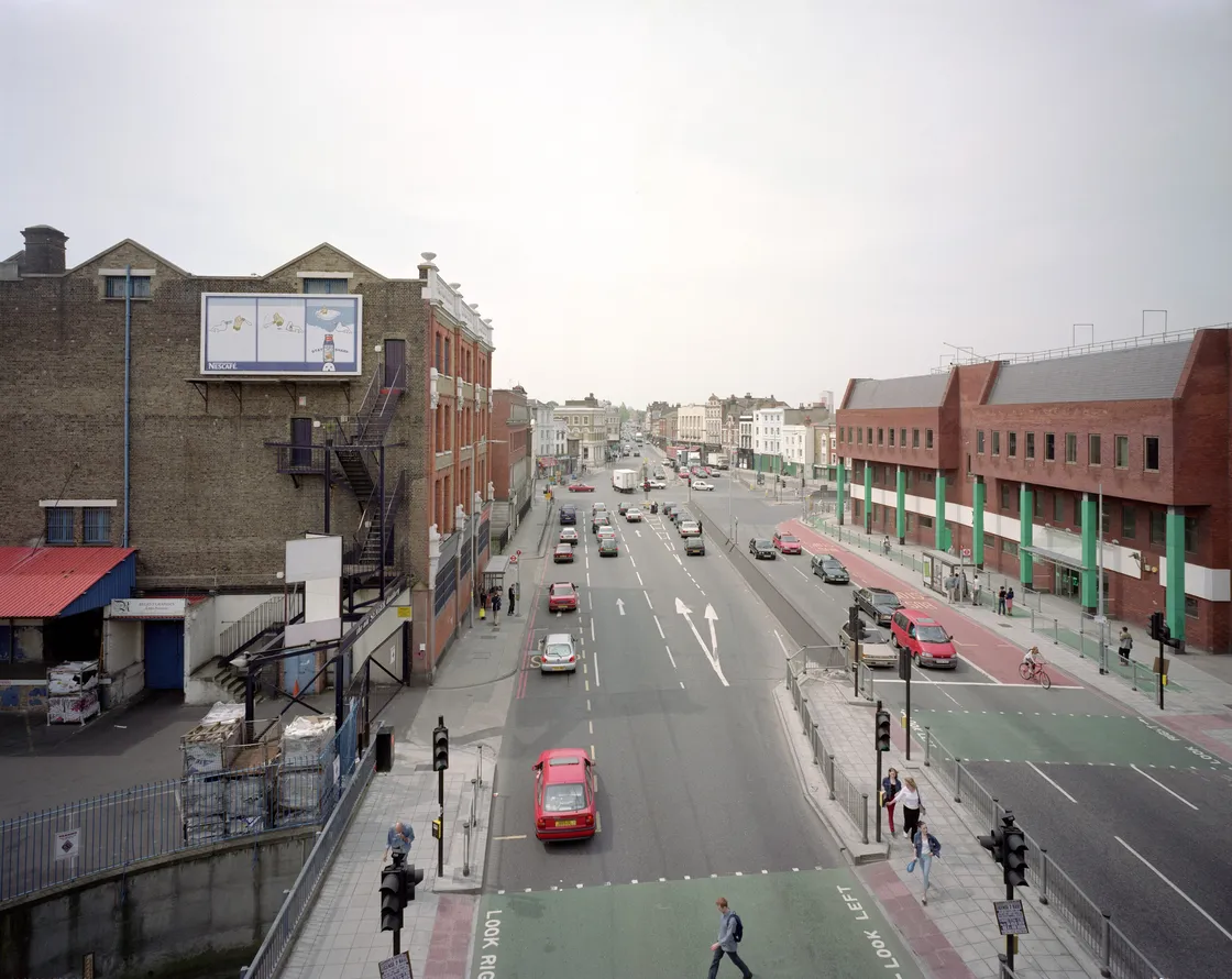 Urban street view showing a road with moderate traffic, bordered by mixed commercial and industrial buildings under a cloudy sky.