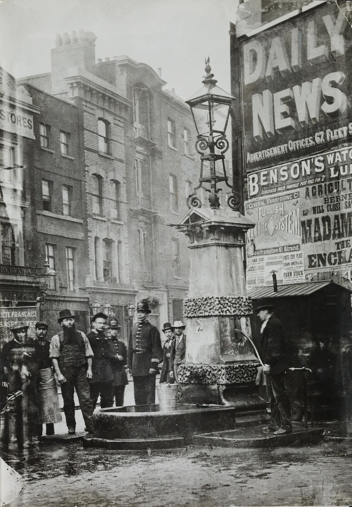 Historical black-and-white photo of people standing around a water fountain on a busy street corner with advertisements.