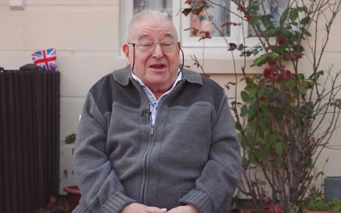 Elderly man in glasses smiling while sitting in a garden with a British flag in the background.