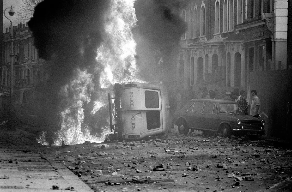 A police van lies overturned and burning on a debris-strewn street with buildings in the background, as a car and a group of people stand nearby.