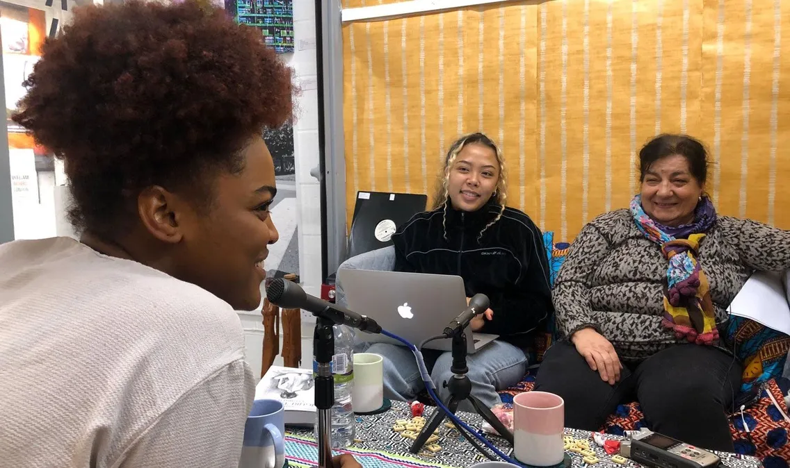 Three women of diverse backgrounds sitting around a table recording a podcast in a cozy, colorful room.