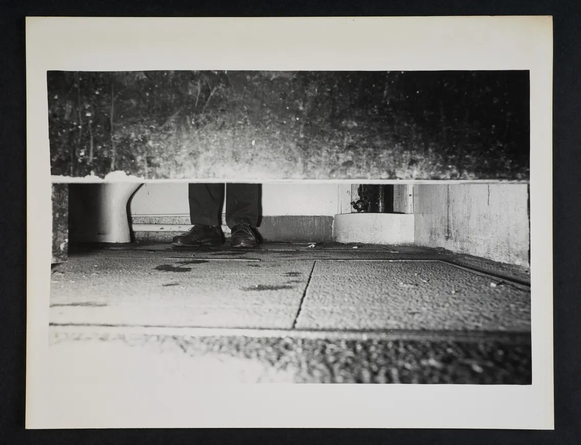 View of the lower part of a closed bathroom stall, showing the feet of a person standing inside. The floor appears wet and slightly dirty.