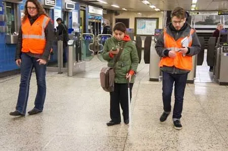 People with visual impairments navigate a subway station, using white canes and mobile devices, accompanied by guides wearing orange vests.