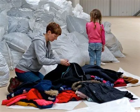 A person sorts through donated clothes while a child stands nearby. Large bags are stacked in the background.