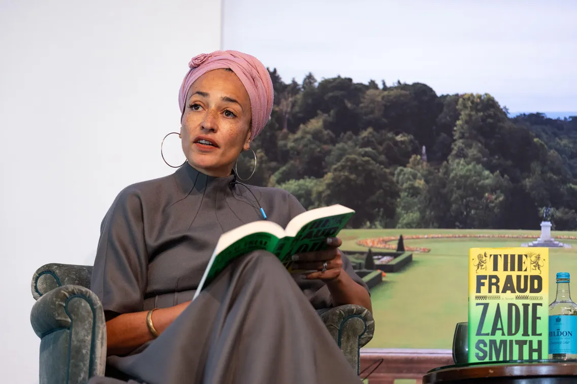 A person wearing a pink headwrap reads from "The Fraud" by Zadie Smith, seated in front of a scenic outdoor backdrop.